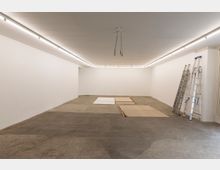 Leerer Raum mit weiß gestrichenen Wänden, einer Decke mit durchgehender Deckenbeleuchtung und einem Betonboden. Am rechten Rand stehen Leitern, und auf dem Boden sind flache Holz- und Leinwandrahmen verteilt.