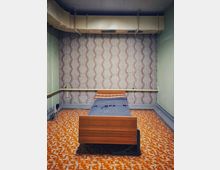 Kleiner, spartanischer Schlafraum mit Einzelbett aus Holz vor einer gemusterten Tapetenwand. Auffällig sind der orange-braun gemusterte Linoleumboden, grün gestrichene Seitenwände sowie große Lüftungs- und Rohrleitungen unter der Decke.