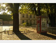 Pflasterplatz mit Bäumen im Vordergrund, einer roten Telefonzelle auf der rechten Seite und einem hellen, historischen Gebäude im Hintergrund. Die Szene ist von warmem Sonnenlicht durchflutet, das durch die Baumkronen fällt.