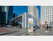 Blick auf den Potsdamer Platz in Berlin mit modernen Gebäuden im Hintergrund und einem Teil der Berliner Mauer im Vordergrund, der mit bunten Graffiti bemalt ist. Menschen versammeln sich auf dem Platz, nahe dem Eingang zur S-Bahn-Station „Bahnhof Potsdamer Platz“.