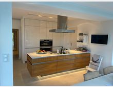 Moderne Küche mit einer zentralen Kochinsel aus Holz und weißer Arbeitsplatte, darüber eine Edelstahl-Dunstabzugshaube. Im Hintergrund ist eine Einbauwand mit hochglänzenden weißen Schränken und integrierten Elektrogeräten, rechts an der Wand befinden sich ein Flachbildfernseher und offene Regale.