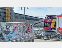 Eine Ansicht der East Side Mall in Berlin mit einem roten Doppeldecker-Hop-on-Hop-off-Bus im Vordergrund. Im unteren Bereich ist eine mit Plakaten und Graffiti bedeckte Wand sowie parkende Fahrräder entlang eines Geländers zu sehen.
