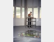 Ein minimalistischer Raum mit großen, halbkreisförmig angeordneten Fenstern, durch die ein Himmel bei Sonnenuntergang sichtbar ist. Im Vordergrund liegt eine transparente Platte mit eingeschlossenen Blumen auf dem Betonboden, während eine Person an einem Metall-Servierwagen arbeitet.