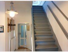 Treppenhaus in einem Wohnhaus mit grauer Holztreppe und weiß gestrichenen Wänden. Links hängt eine Wanddekoration aus Holz neben einer weißen Tür mit Glaseinsätzen; darüber ist eine einfache Deckenlampe mit einer großen Glühbirne montiert.