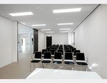 Ein moderner Seminarraum mit mehreren schwarzen Stühlen in Reihen angeordnet, helle Wände, eine abgehängte Decke mit integrierter Beleuchtung und ein Fenster rechts. Im Hintergrund führt eine offene Schiebetür zu einem angrenzenden Raum.