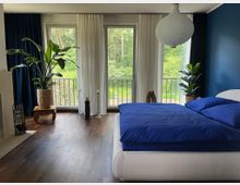 Modernes Schlafzimmer mit dunklem Holzboden, blau gestrichener Akzentwand und großem Bett mit blauer Bettwäsche. Drei bodentiefe Fenster mit weißen Vorhängen öffnen den Blick auf einen Balkon und viel Grün; große Zimmerpflanzen und eine runde Papier-Hängelampe ergänzen die Einrichtung.