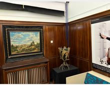 Innenraum mit dunkler Holzvertäfelung und weißer Decke, in dem ein gerahmtes Landschaftsgemälde über einem Heizkörper hängt. Daneben steht auf einem schwarz bedeckten Tisch eine skulpturale Geweih-Installation, rechts ist ein großes Wandposter zu sehen.