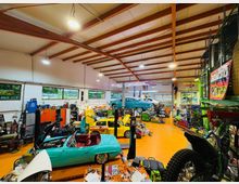 Große Autowerkstatt mit hohen Deckenbalken, orangefarbenem Boden und vielen Werkbänken, Regalen sowie Hebebühnen. Mehrere Oldtimer und Fahrzeuge stehen im Innenraum, teils mit geöffneten Motorhauben, neben Werkzeug und Ersatzteilen.