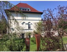 Historisches Gebäude mit hellem Putz, rotem Satteldach und Arkadenfenstern, umgeben von einem Garten mit blühenden Bäumen und einigen rostigen Metallskulpturen im Vordergrund. Der Himmel ist klar und blau.