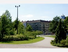 Städtischer Park mit Rasenflächen, Büschen und Bäumen im Vordergrund, einem gepflasterten Weg und einer Straßenlaterne. Im Hintergrund befinden sich Wohngebäude mit mehreren Etagen und Ziegeldächern.