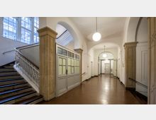 Treppenhaus und Flur eines historischen Gebäudes mit hohen Decken und einer Kombination aus weiß gestrichenen Wänden und Sandsteinsäulen. Auf der linken Seite befindet sich eine Treppe mit verziertem Metallgeländer, während der Flur geradeaus durch einen Rundbogen verläuft, beleuchtet von hängenden Kugelleuchten.