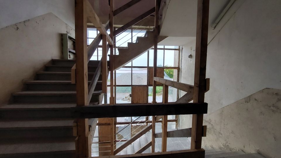 Treppenhaus in einem unvollendeten oder renovierungsbedürftigen Gebäude mit sichtbar rohen Holzgeländern und großen Fenstern, die nach außen auf eine Kiesfläche und Vegetation blicken. Die Wände sind weiß und teilweise beschädigt.