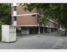 Wohnhaus aus rotem Backstein mit erhöhtem Erdgeschoss auf Pfeilern, flankiert von Bäumen auf einer gepflasterten Straße. Im Vordergrund steht ein Altkleidercontainer, im Hintergrund sind weitere Gebäude und parkende Autos zu sehen.