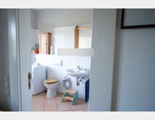 Blick durch eine Tür in ein kleines Badezimmer mit weißen Fliesen, rosa Bodenfliesen, WC und Waschbecken. An der Wand hängen ein Spiegel und ein kleiner Holzschrank, daneben steht ein Tritthocker und ein Heizkörper.