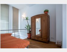 Ein Schlafzimmer mit Holzboden, einem großen Schrank mit antikem Design und Spiegel in der Tür, sowie einer Zimmerpflanze in einem Topf neben dem Schrank. Im Vordergrund befindet sich ein Bett mit orangefarbener Bettwäsche, während durch hohe Fenster mit Jalousien Tageslicht einfällt.