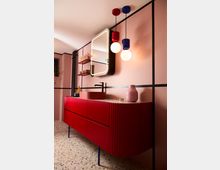 Modernes Badezimmer mit rosa Wänden und geometrischen schwarzen Linien. Im Vordergrund steht ein markantes rotes Waschbeckenmöbel mit Rippenstruktur, darüber ein beleuchteter Spiegel; farbige Hängelampen in Rot und Blau ergänzen das Design, der Boden ist mit Terrazzo-Fliesen gestaltet.