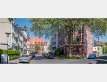 Straßenszene mit historischen Wohngebäuden, darunter ein rotes Eckhaus mit weißen Verzierungen rechts, und einem roten Ziegelgebäude mit Spitzdach im Hintergrund. Die Straße ist von Autos gesäumt und von einem großen Baum überschattet.