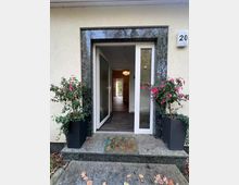 Eingang eines Wohnhauses mit geöffneter weißer Tür und seitlichem Glaselement, eingefasst von dunklem Stein. Vor der Schwelle liegen eine Fußmatte und eine kleine Stufe; links und rechts stehen große Pflanzkübel mit blühenden Sträuchern, und im Flur ist eine Deckenleuchte sichtbar.