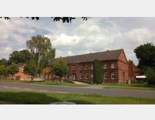 Backsteingebäude im ländlichen Stil mit zwei Stockwerken, weißen Sprossenfenstern und einem Ziegeldach, umgeben von Bäumen und Rasenflächen. Im Vordergrund verläuft eine zweispurige Straße.