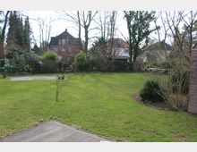 Großer Garten mit gepflegtem Rasen, einzelnen Büschen und jungen Bäumen, umgeben von Bäumen und Hecken. Im Hintergrund sind Wohnhäuser und im Vordergrund eine Holzterrasse sichtbar.