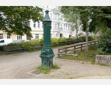 Kleiner Stadtpark mit einem dekorativen, grünen Trinkwasserbrunnen aus Metall im Vordergrund, Sitzbänken entlang der Wege und Bäumen sowie Sträuchern, im Hintergrund mehrstöckige Wohnhäuser mit weißen Fassaden.