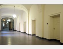 Ein heller Flur mit hohen gewölbten Decken, cremefarbenen Wänden und mehreren Türöffnungen auf der rechten Seite. Am Ende des Flurs befindet sich eine verzierte grüne Doppeltür aus Metall mit Glasflächen, durch die ein weiterer Raum sichtbar ist; runde Deckenleuchten hängen von der Decke.