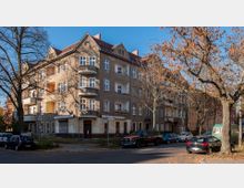 Eckgebäude eines mehrstöckigen Wohnhauses aus der Vorkriegszeit mit Putzfassade, mehreren Balkonen und roten Ziegeldächern. Im Vordergrund stehen geparkte Autos entlang einer von Bäumen gesäumten Straße.