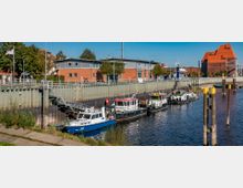 Kleine Hafenszene mit mehreren festgemachten Booten entlang einer Kaimauer vor einer modernen, roten Ziegelarchitektur; im Hintergrund ein großes Industriegebäude mit markantem orangefarbenem Dach.