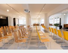 Heller Veranstaltungsraum mit mehreren Reihen Holz- und gelber Kunststoffstühle, die auf ein Podium ausgerichtet sind. Der Raum hat weiße Wände, große Fenster, Vorhänge und vereinzelt Kübelpflanzen.