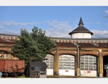 Backstein-Rundlokschuppen mit halbrunden Toren und einem zentralen Kegeldach-Turm; davor stehen ein rostiger Güterwaggon, ein schwarzer Eisenbahnwagen und zwei Nadelbäume.