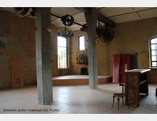 Großer Innenraum einer alten Brennerei mit Backsteinwänden, hohen Fenstern und freistehenden Betonstützen. Im Raum befinden sich ein erhöhtes Podest, antike Maschinen, ein hölzernes Rednerpult und Stühle.