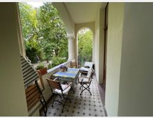 Überdachte Veranda eines historischen Gebäudes mit gemusterten Fliesen, einem Tisch mit Tischdecke und mehreren Stühlen. Im Hintergrund sind große Bäume und eine grüne Gartenlandschaft zu sehen.