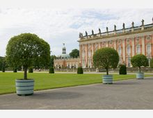 Barockes Schloss mit roten Fassaden und weißen Fensterelementen, flankiert von Statuen auf dem Dach. Im Vordergrund stehen symmetrisch angeordnete Bäume in runden Pflanzkübeln auf einem gepflegten Rasen mit gepflasterten Wegen.