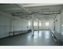 Ein leerer, industrieller Raum mit weißen Wänden und Fliesen, ausgestattet mit langen Edelstahltischen entlang der Seitenwände. Der Raum hat mehrere Fenster, säulenartige Stützen und an der Decke verlaufen freiliegende Kabel und Rohre.