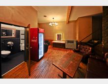 Ein Raum mit Holzboden, einer roten Kühlschrankbox mit der Aufschrift "ASTRA", einer Spiegelkonsole mit Beleuchtung, braunen Ledersofas und einem rustikalen Holztisch. Im linken Bereich ist ein angrenzendes Badezimmer mit dunklen Fliesen und einer Toilette sichtbar.