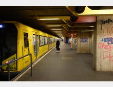Eine U-Bahn-Station mit einem gelben Zug an der linken Seite des Bahnsteigs. Die Wände sind aus Beton und mit Graffiti bedeckt, während die gelb-schwarzen Deckenstrukturen Leuchten enthalten; eine Person steht auf dem Bahnsteig.