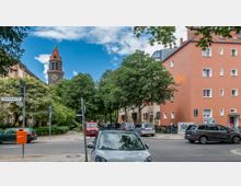 Straßenszene in einem Wohngebiet mit mehrstöckigen Wohnhäusern, einem markanten Kirchturm aus Backstein mit Uhr im Hintergrund und Bäumen entlang der Straße. Im Vordergrund parken mehrere Autos, und ein Straßenschild markiert die „Lauenburger Str.“.