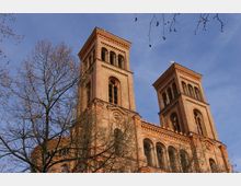 Backstein-Kirche mit zwei hohen Türmen und Rundbogenfenstern, umgeben von kahlen Ästen eines Baumes vor einem klaren, blauen Himmel.
