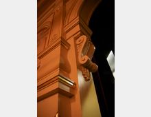Detailaufnahme eines architektonischen Elements in einem Gebäude, mit verzierten Pilastern, geschwungenen Ornamenten und goldenen Akzenten. Der Hintergrund zeigt einen dunklen Bühnenraum mit einem roten Vorhang.