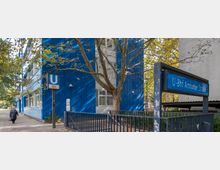 Außenansicht der U-Bahn-Station "U-Bhf Amrumer Straße" in Berlin, mit einem blauen Stationsschild und einem Gebäude mit blauer Fassade im Hintergrund. Ein Baum steht zentral im Bild, umgeben von Gehweg und einem Geländer.