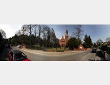 Panoramaaufnahme einer roten Backsteinkirche im neugotischen Stil mit hohem Turm und Spitzdach, umgeben von einem umzäunten Garten mit Bäumen. Die Kirche steht an einer ruhigen Straße mit geparkten Autos, flankiert von Wohnhäusern und Bäumen.