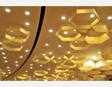 Beleuchtete Decke eines modernen Gebäudes mit sechseckigen, goldfarbenen Hängeleuchten und zahlreichen runden Einbauspots, die auf einer perforierten Deckenstruktur montiert sind.