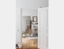 Ein kleines Badezimmer mit Holzboden und weiß gestrichenen Wänden, ausgestattet mit einem Waschbecken, einem rechteckigen Spiegel in einem Holzrahmen, einem Korbstuhl mit braunem Handtuch und einer Pflanze im Hintergrund. Im Vordergrund steht eine geöffnete weiße Tür mit verzierten Paneelen.