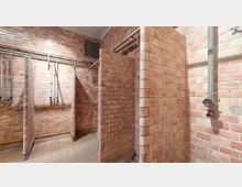 Duschraum mit Wänden aus rötlichem Ziegelmauerwerk, mehreren offenen Duschkabinen und freiliegenden Rohren an der Decke. Der Boden ist mit hellen Fliesen ausgestattet, und die Duschbereiche haben einfache Trennwände aus Ziegeln.