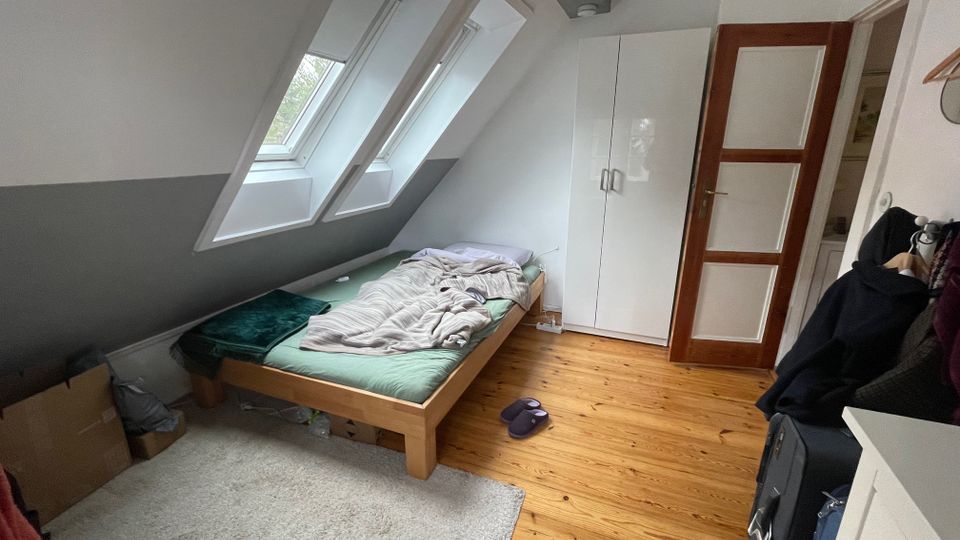 Kleines Dachgeschoss-Schlafzimmer mit schrägen Wänden, zwei großen Dachfenstern und Holzboden. Im Raum stehen ein niedriges Bett, ein weißer Kleiderschrank und eine Holztür mit Glaseinsätzen.