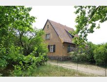 Ein kleines, historisches Wohnhaus mit gelber Fassade, schwarzen Fensterläden und einem roten Ziegeldach, umgeben von dichtem Grün, Sträuchern und Bäumen. Ein schmaler, unbefestigter Weg führt an der Seite des Hauses vorbei.