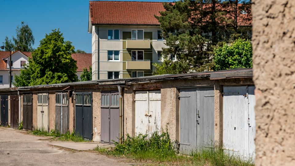 Reihe älterer Garagen mit unterschiedlich gestrichenen Holztoren entlang einer asphaltierten Zufahrt, teils mit Bewuchs am Boden. Dahinter stehen mehrgeschossige Wohnhäuser mit roten Ziegeldächern und Bäumen unter blauem Himmel.