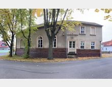 Eckgebäude mit verputzter Fassade und Klinkersockel, umgeben von Bäumen mit herbstlichem Laub. Das Gebäude hat weiß gerahmte Fenster, Rundbogenfenster auf der linken Seite und einen kleinen Eingang mit Treppe.