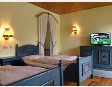 Ein traditionell eingerichtetes Schlafzimmer mit zwei Einzelbetten aus Holz, blauen Möbeln mit Blumenmotiven, einer Kommode mit Fernseher und gelben Wänden. Im Hintergrund ist ein Durchgang mit weißen Vorhängen zu sehen, die Holzdecke und Holzfußboden ergänzen den rustikalen Stil.