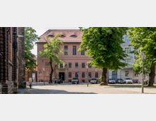 Ein Platz mit Kopfsteinpflaster, flankiert von einem historischen Backsteingebäude links und großen Bäumen. Im Zentrum steht ein zweistöckiges Gebäude mit rosa Fassade, einem Eingangsportal aus Stein und einem roten Ziegeldach; im Hintergrund sind parkende Autos zu sehen.